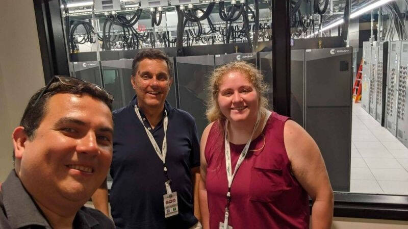 Three people wearing ID badges stand in front of a window, behind which are visible server racks and cables in a high-tech data center environment.